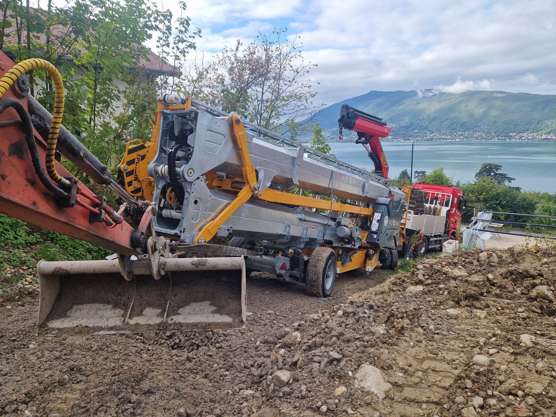 Une excavatrice et un camion-pompe à béton se trouvent à flanc de colline, avec vue sur un lac et des montagnes.
