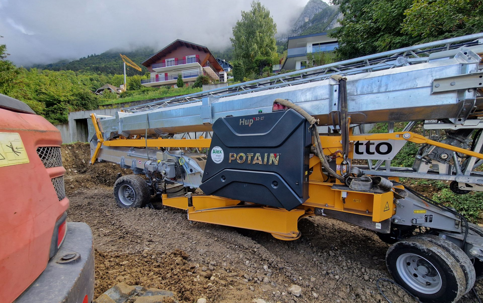 Une grue jaune Potain sur une route boueuse, avec des montagnes et des bâtiments en arrière-plan.
