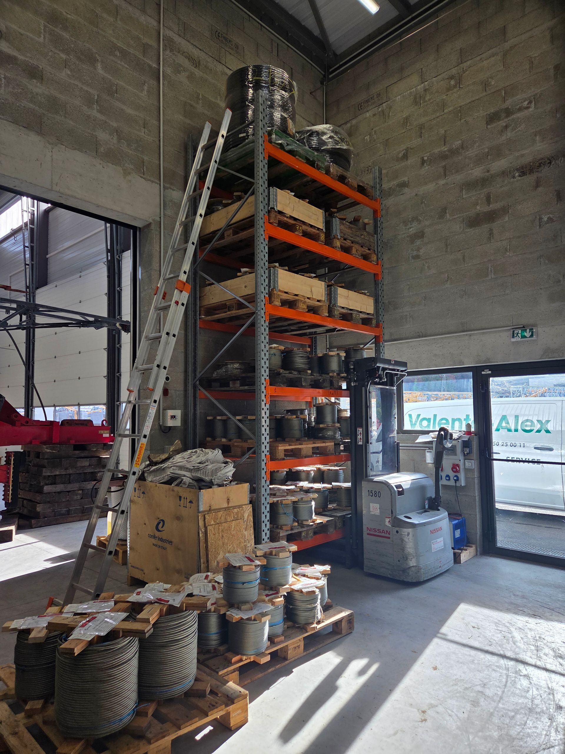 Intérieur de l'entrepôt : étagères avec des marchandises emballées, échelle, bobines de fil et chariot élévateur.