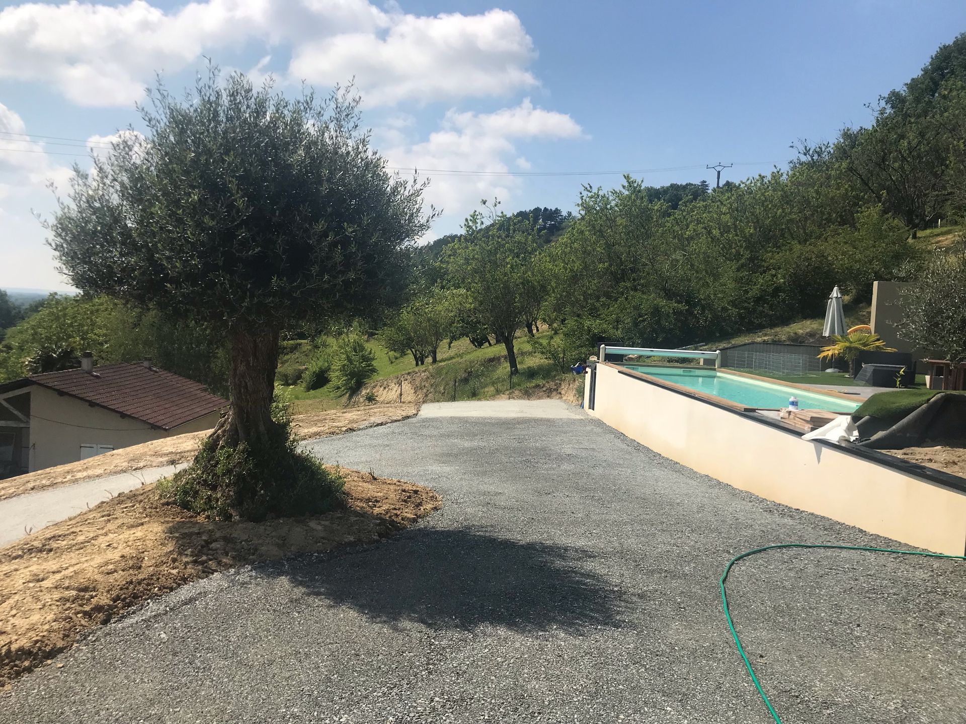 Allée de gravier bordée d'un arbre menant à une piscine offrant une vue sur les arbres et un ciel d'un bleu éclatant.