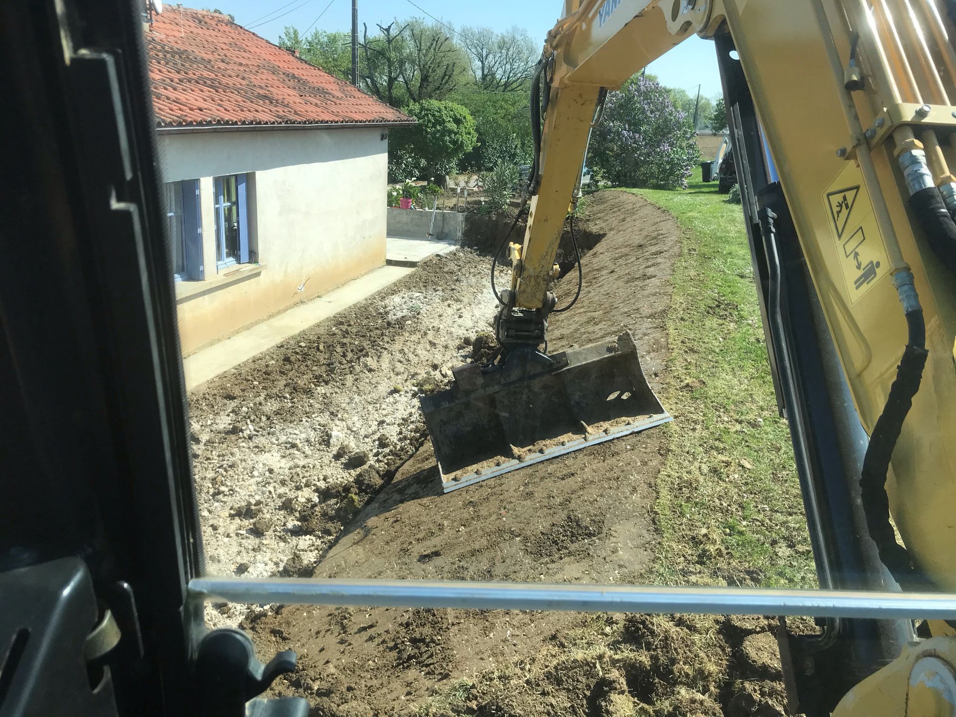 Une pelleteuse creuse une tranchée à côté d'une maison au toit de tuiles rouges.