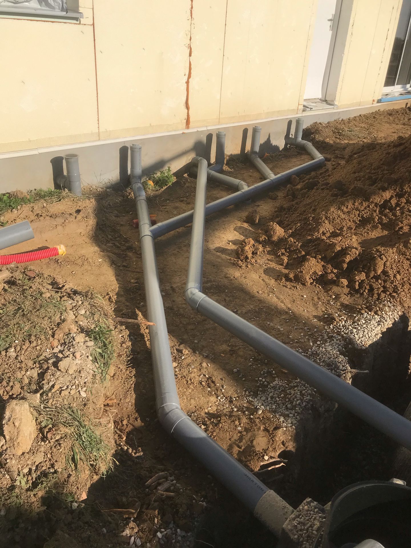 Des tuyaux en PVC gris sont installés dans une tranchée le long des fondations d'un bâtiment.