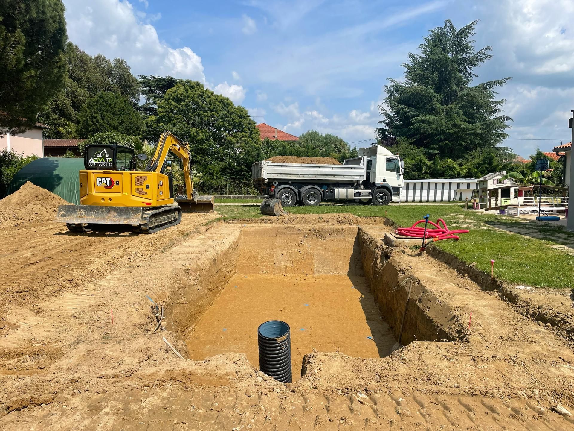 Chantier de fouilles avec une excavatrice jaune, un camion-benne et une fosse rectangulaire.