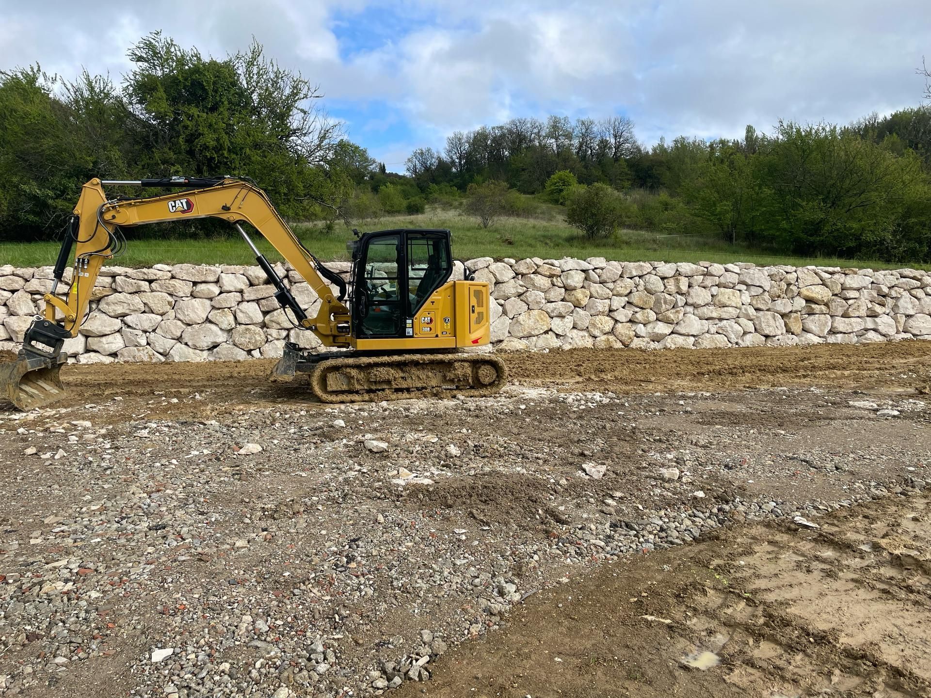 Une pelleteuse jaune déblaye du gravier sur un terrain meuble, avec un mur de soutènement en pierre en arrière-plan.