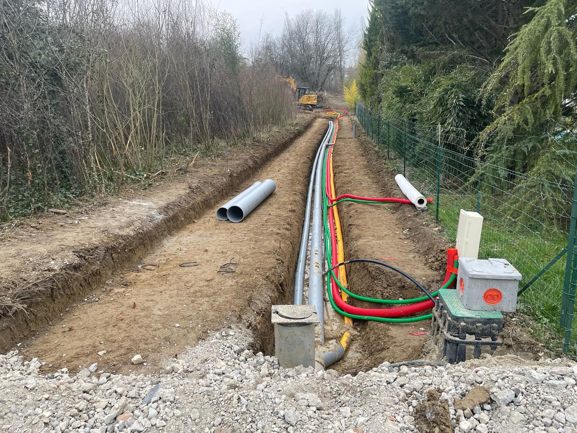 Tranchée de construction avec conduits, câbles et gravier, à proximité d'une clôture et de végétation.