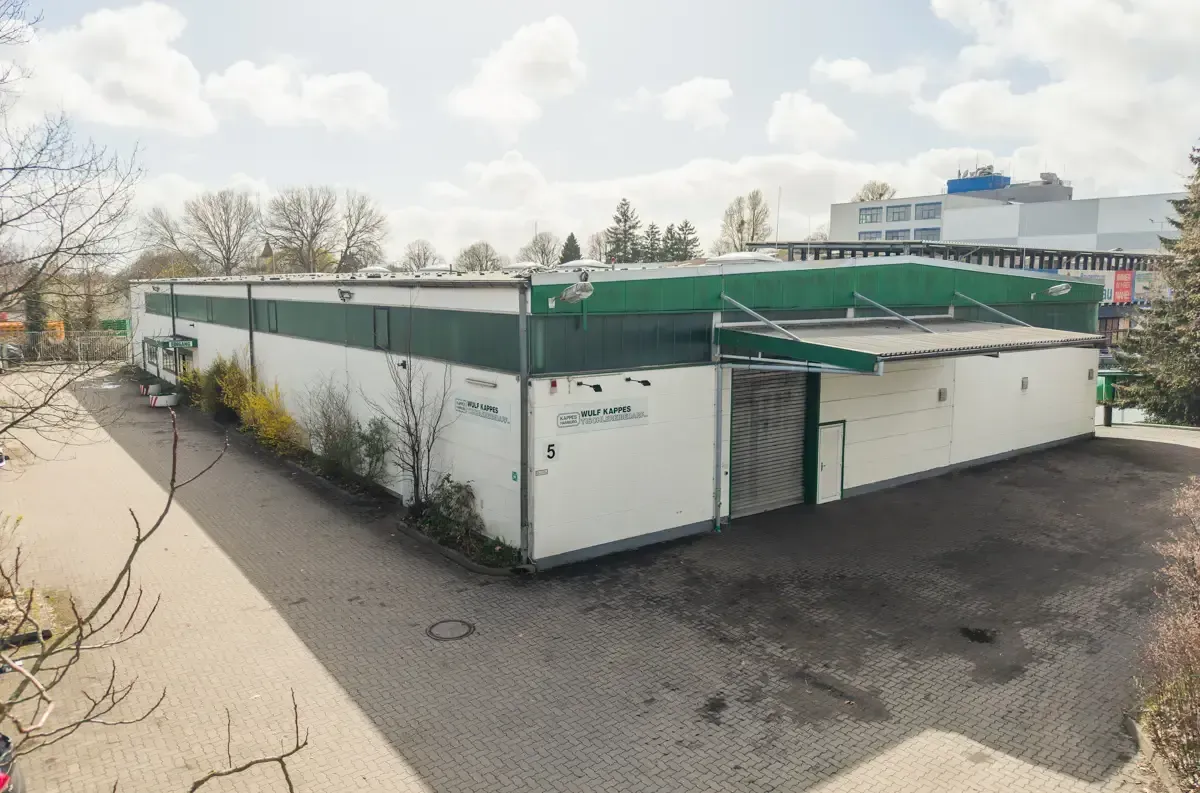 Weißes Industriegebäude mit Gründach neben einem gepflasterten Parkplatz und Bäumen unter einem bewölkten Himmel