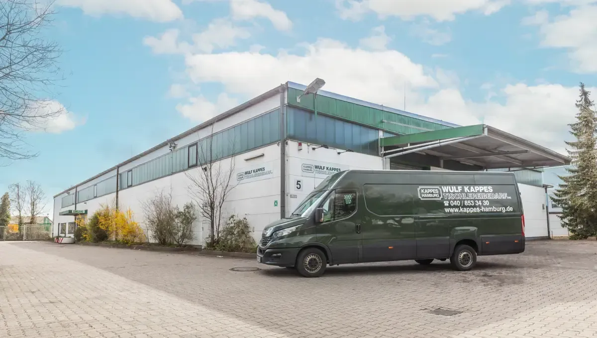 Ein dunkelgrüner Lieferwagen parkt neben einem weißen Industriegebäude mit Glasdach unter blauem Himmel.