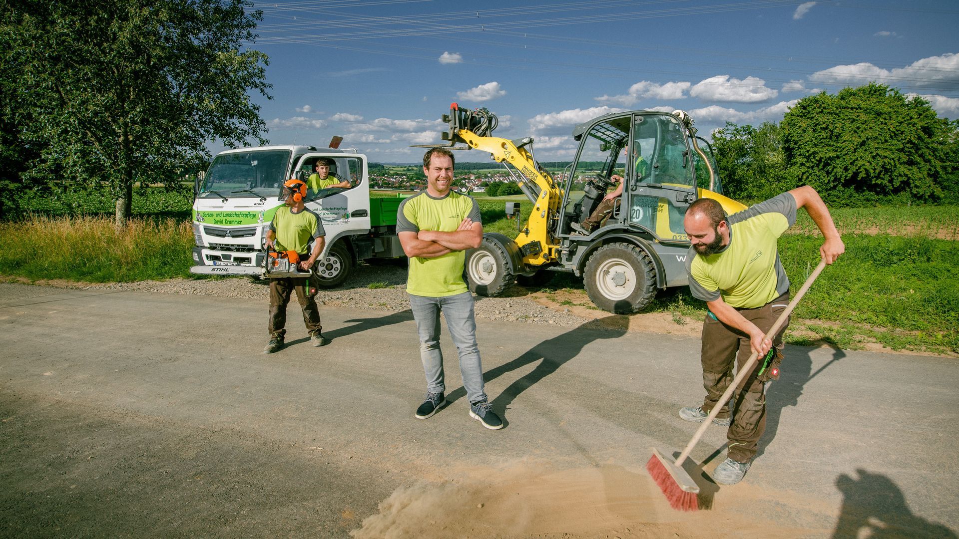 Arbeiter in Warnwesten reparieren mit einem Bagger und einer Schaufel einen Graben am Straßenrand in einem ländlichen Gebiet.