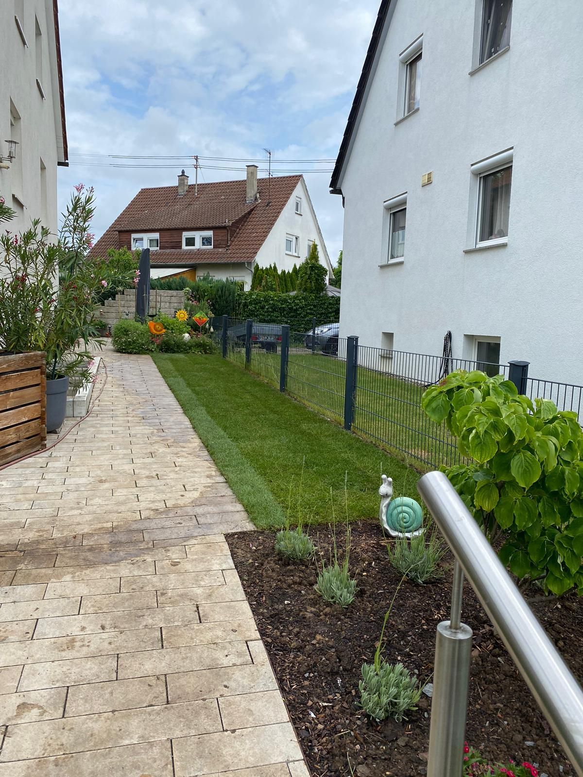 Schmaler Gartenweg neben weißen Häusern, mit Rasen, Blumenbeeten und bewölktem Himmel.