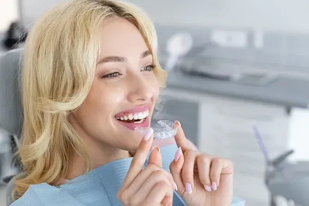 Una mujer está sentada en una silla dental sosteniendo un aparato transparente en su boca.