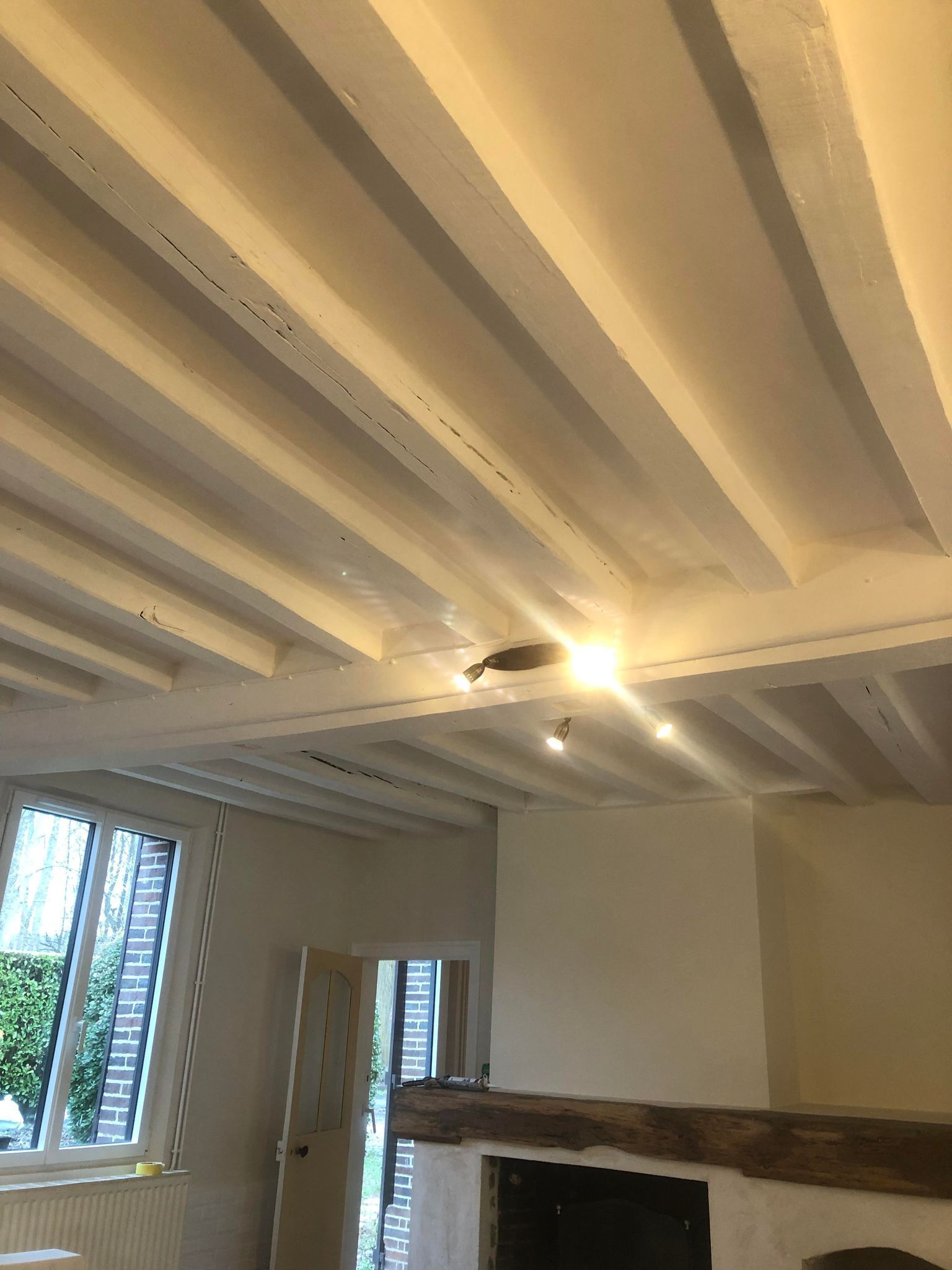 Plafond à poutres apparentes peint en blanc dans une pièce avec cheminée et fenêtre.