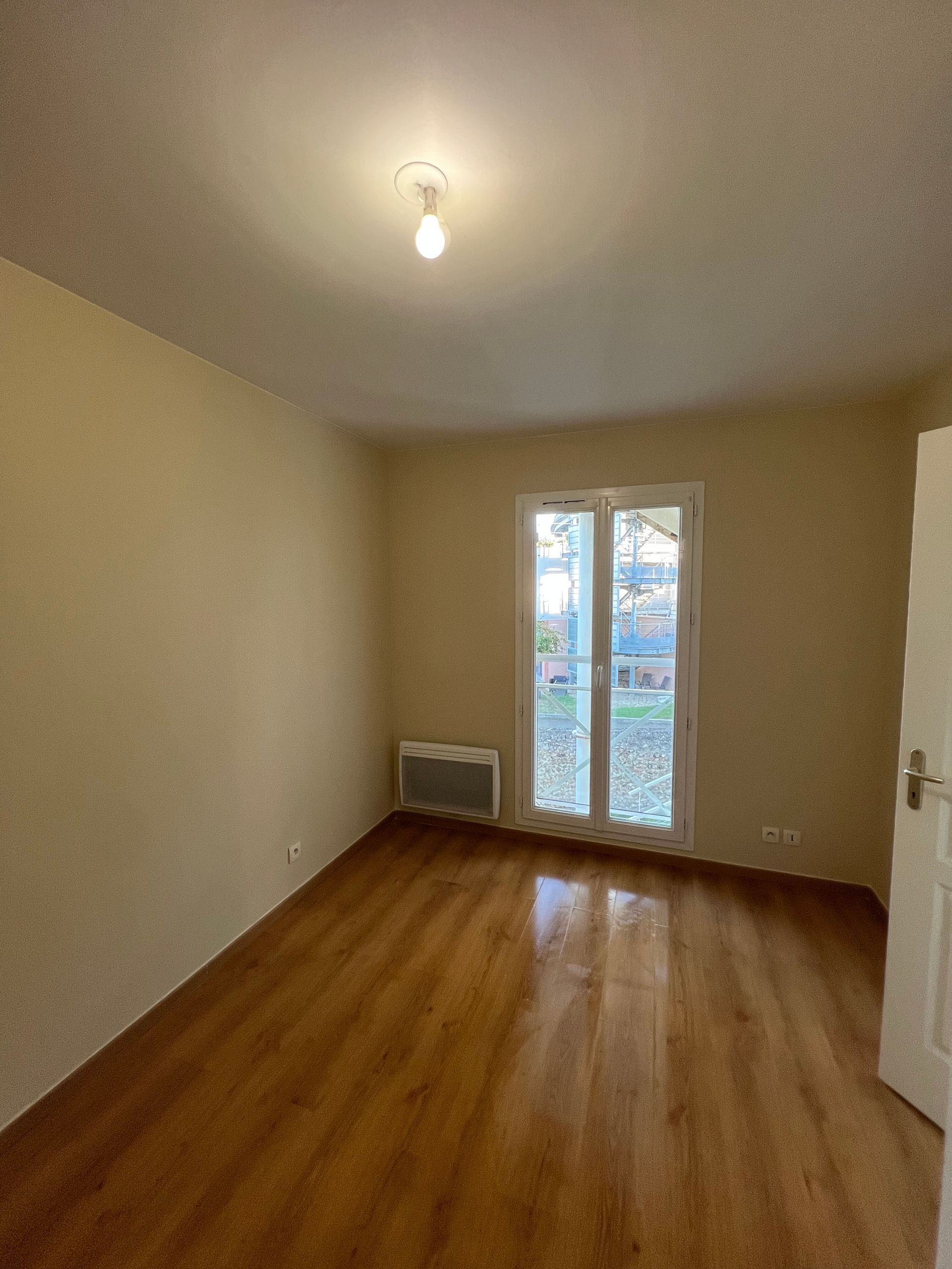 Pièce vide avec parquet, une porte et une fenêtre donnant sur la verdure. Murs beiges, éclairage au plafond.