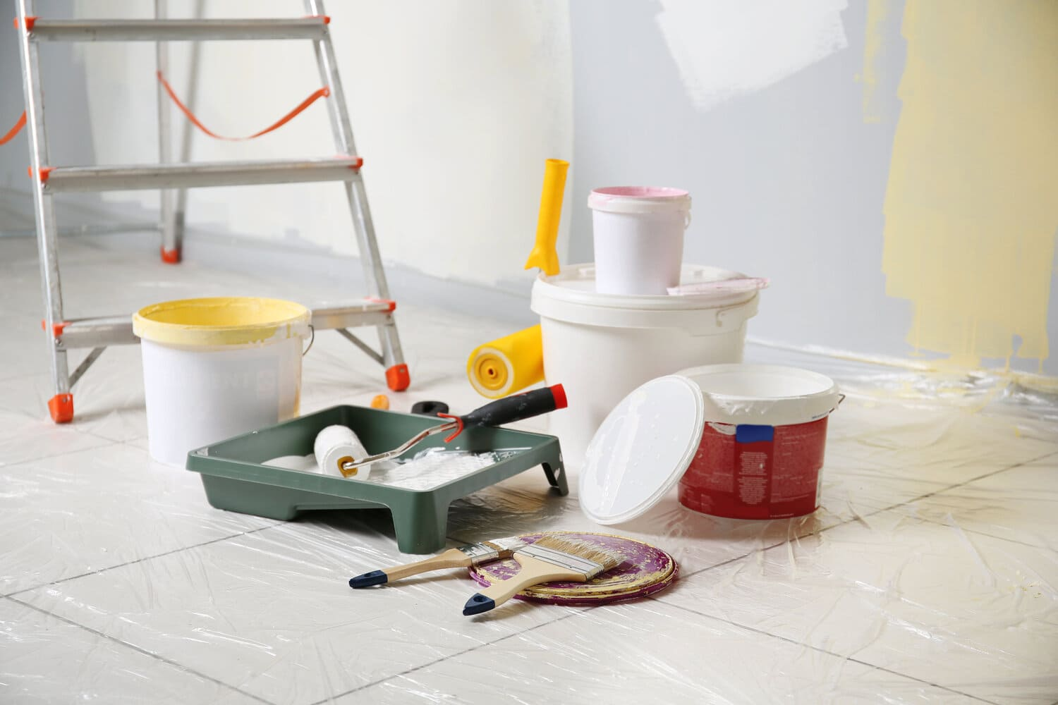 En una habitación que se está pintando hay cubos de pintura, rodillos, pinceles y una escalera.