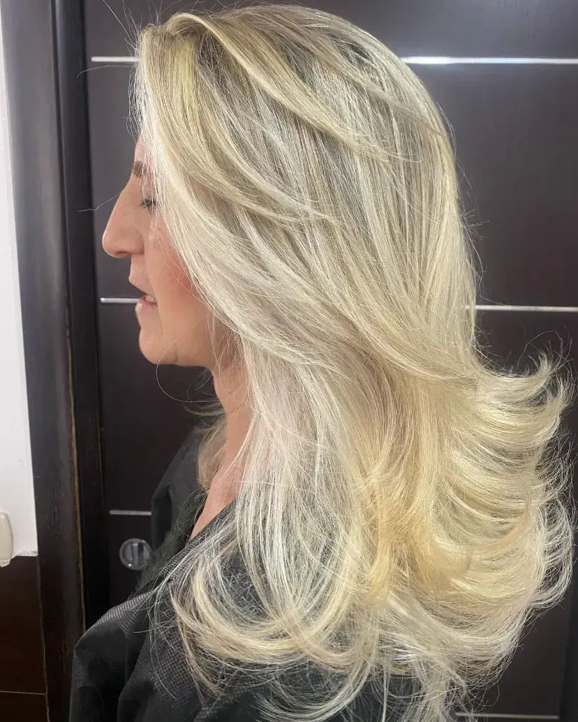 Mujer con cabello rubio largo en capas, vista de perfil lateral, fondo oscuro.