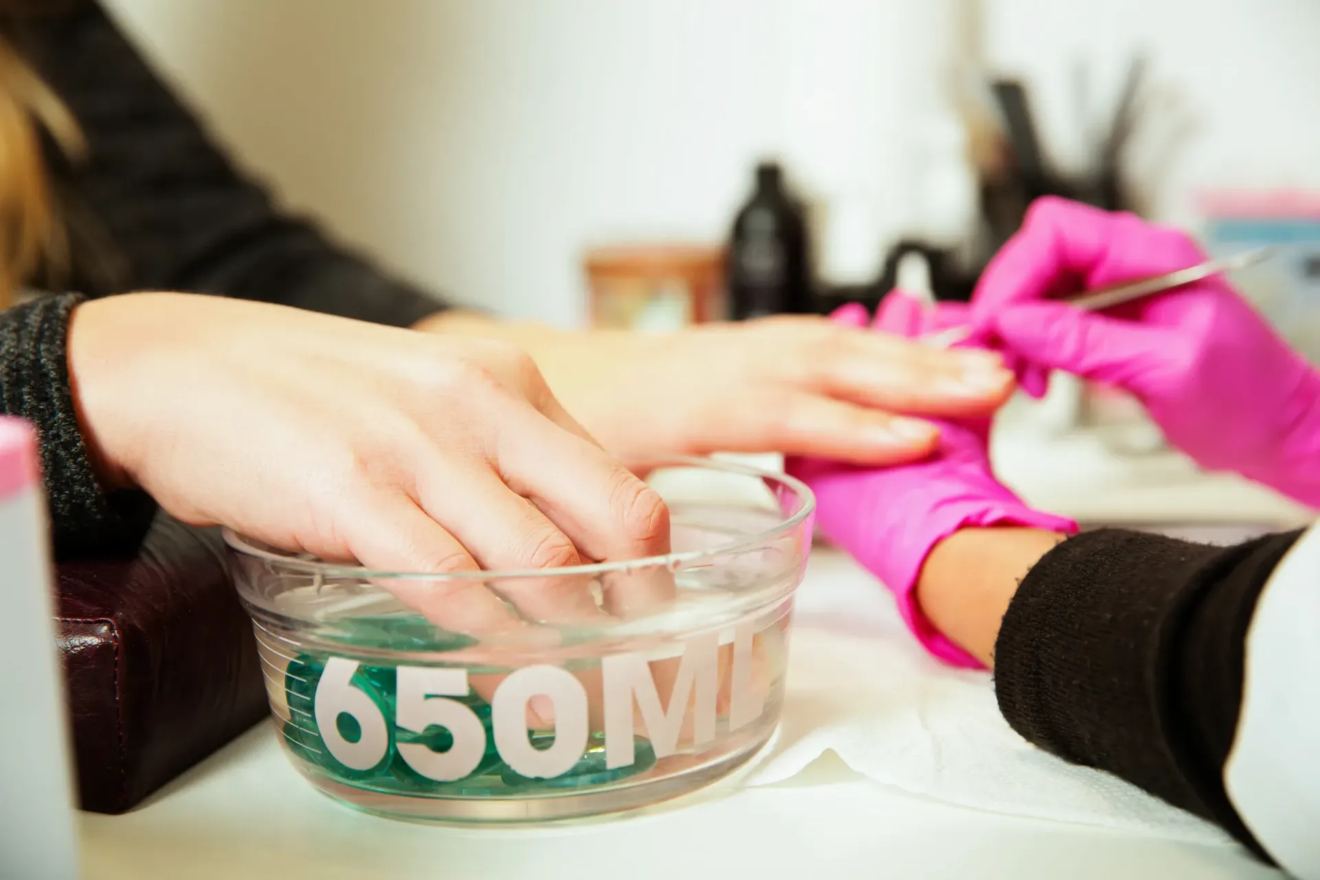 La mano de una persona se remoja en un recipiente etiquetado, mientras otra persona con guantes rosas se prepara las uñas.