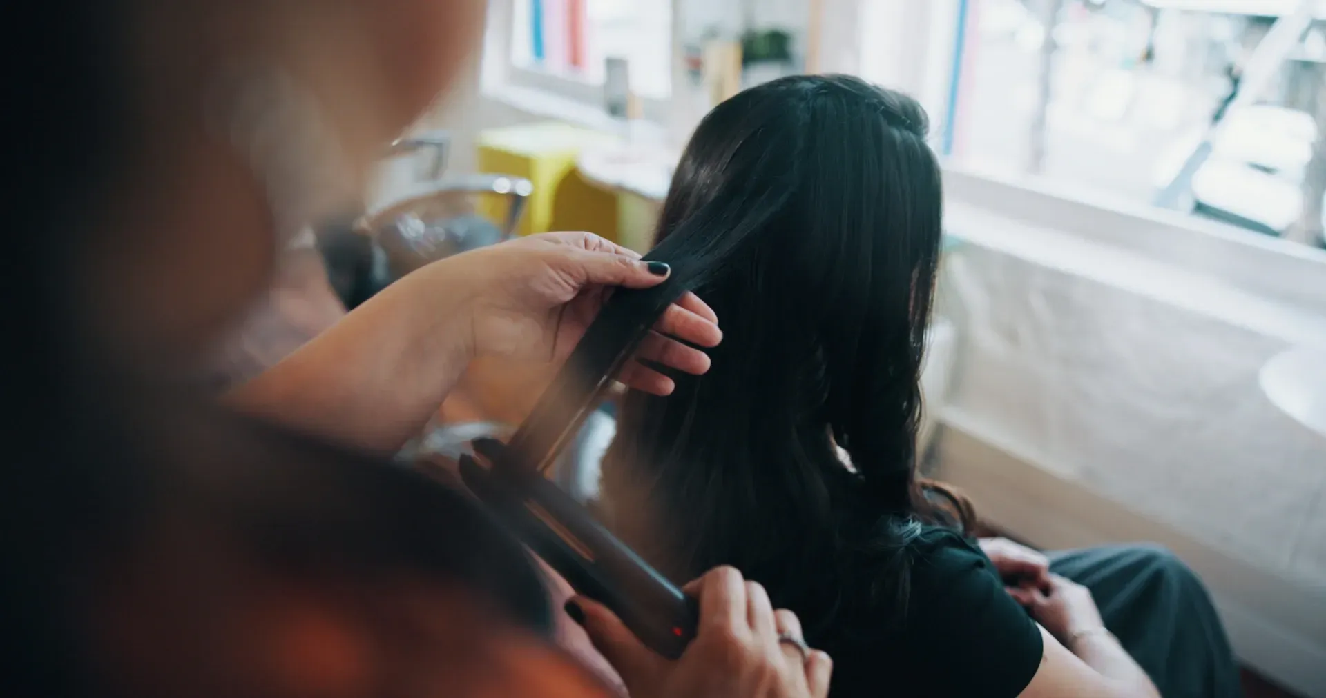 Un peluquero riza el cabello largo y oscuro de una clienta con una plancha en un salón, cerca de una ventana.