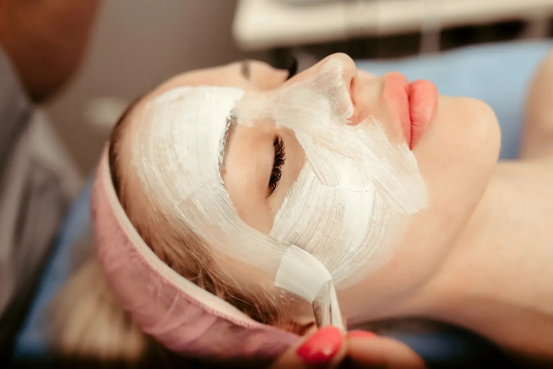 Persona recibiendo tratamiento facial: mascarilla blanca aplicada con pincel, diadema rosa, acostado.