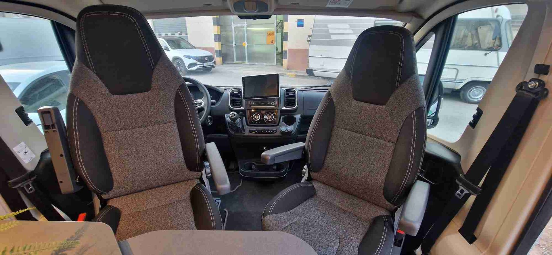 Vista interior de una autocaravana con dos asientos delanteros mirando hacia el salpicadero. Color neutro; vehículo estacionado.