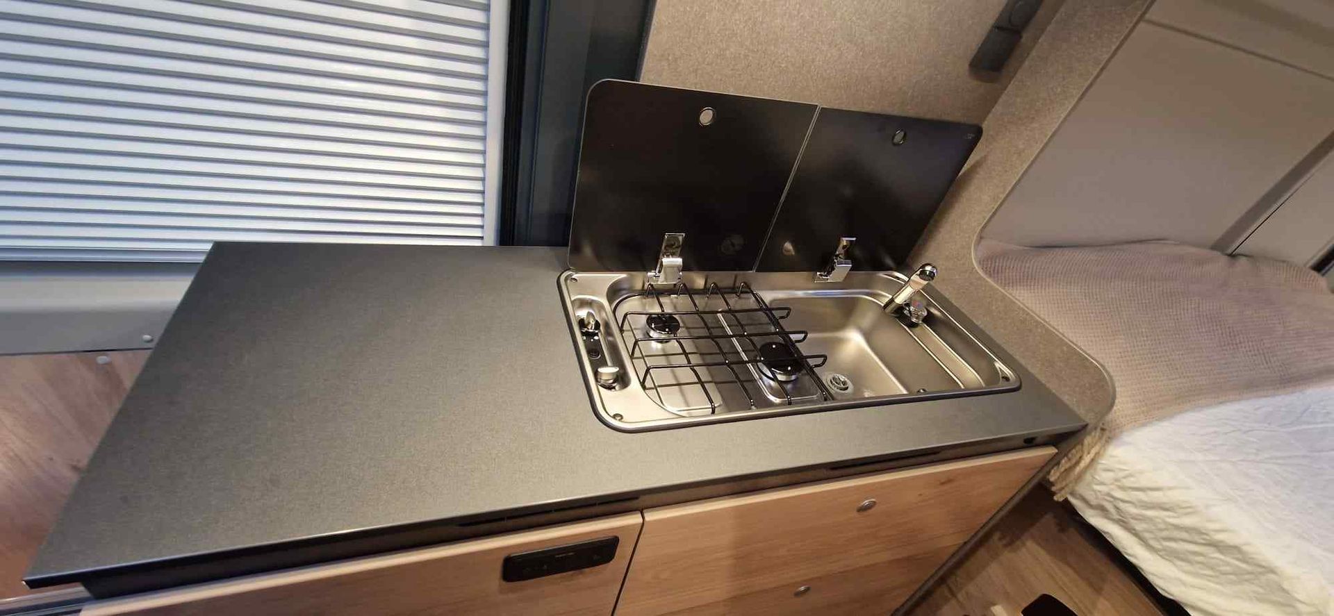 Interior de la cocina de una furgoneta camper con estufa, fregadero, encimera y una cama.