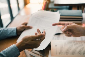 Dos personas revisando documentos, una señalando con un bolígrafo. Un escritorio con libros.