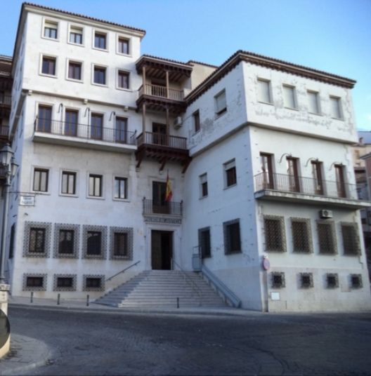 Edificio blanco con balcones y una bandera española sobre la entrada; unos escalones conducen a la puerta.