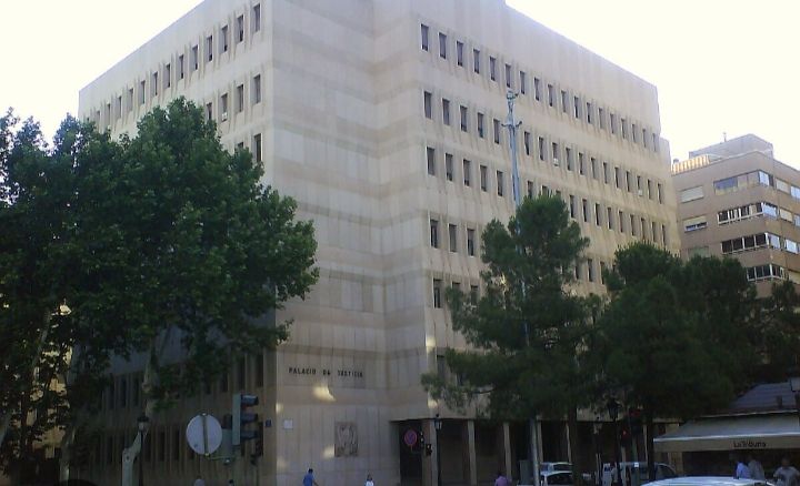 Un gran edificio de piedra con múltiples hileras de ventanas, árboles y un cartel en la calle. El edificio lleva la inscripción "PALAU DE JUSTICIA".