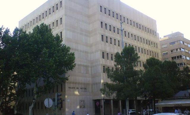 Un gran edificio de piedra con múltiples hileras de ventanas, árboles y un cartel en la calle. El edificio lleva la inscripción "PALAU DE JUSTICIA".