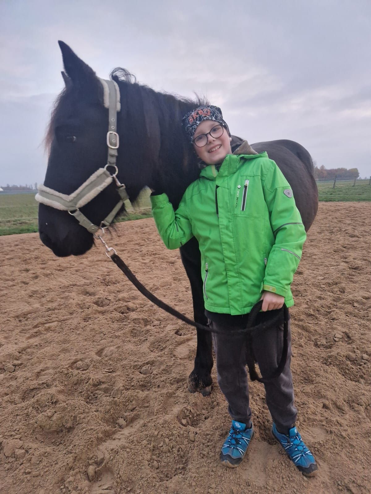 Ein kleiner Junge in einer grünen Jacke steht neben einem schwarzen Pferd.