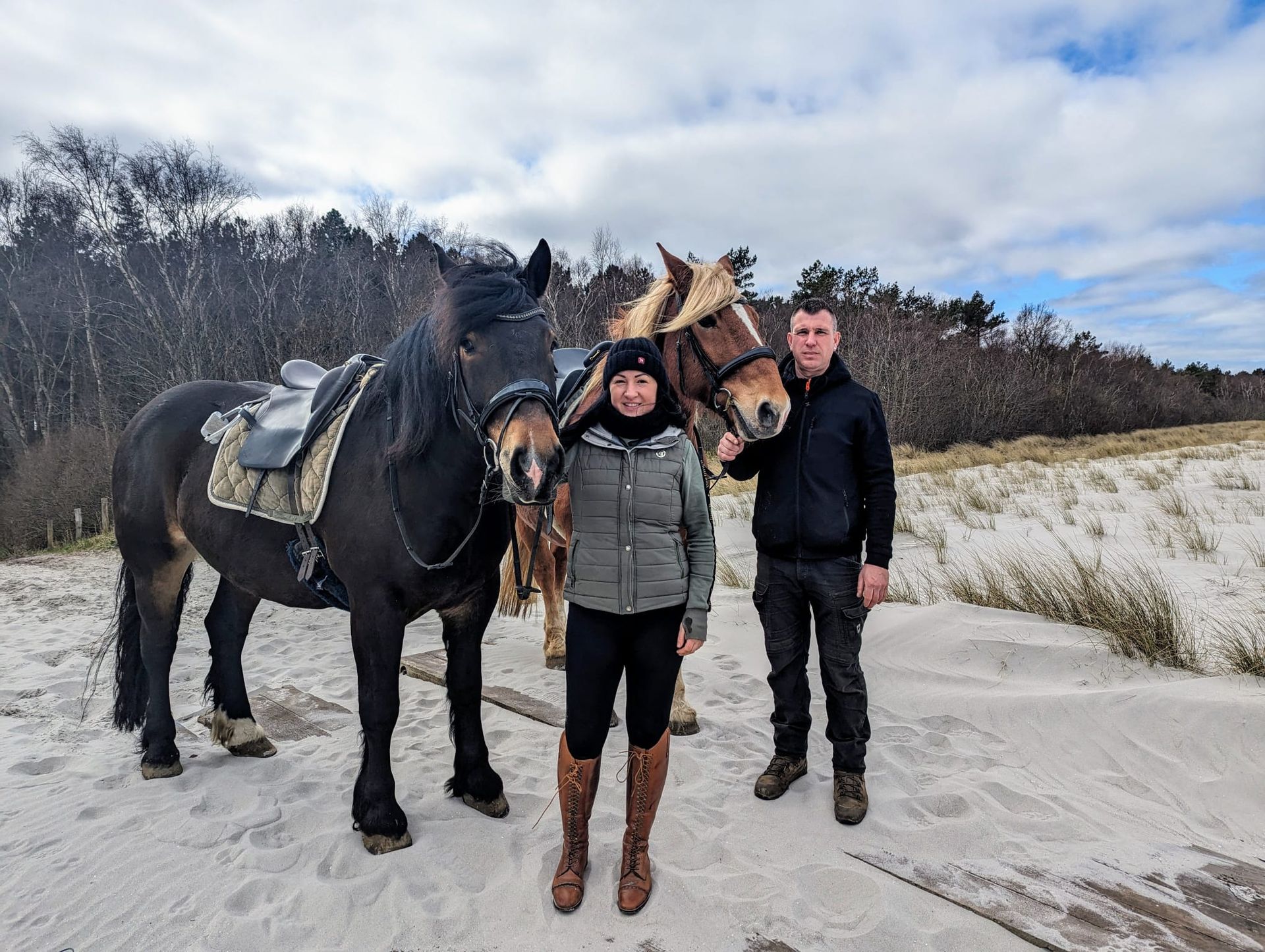 Ein Mann und eine Frau stehen neben zwei Pferden im Sand.