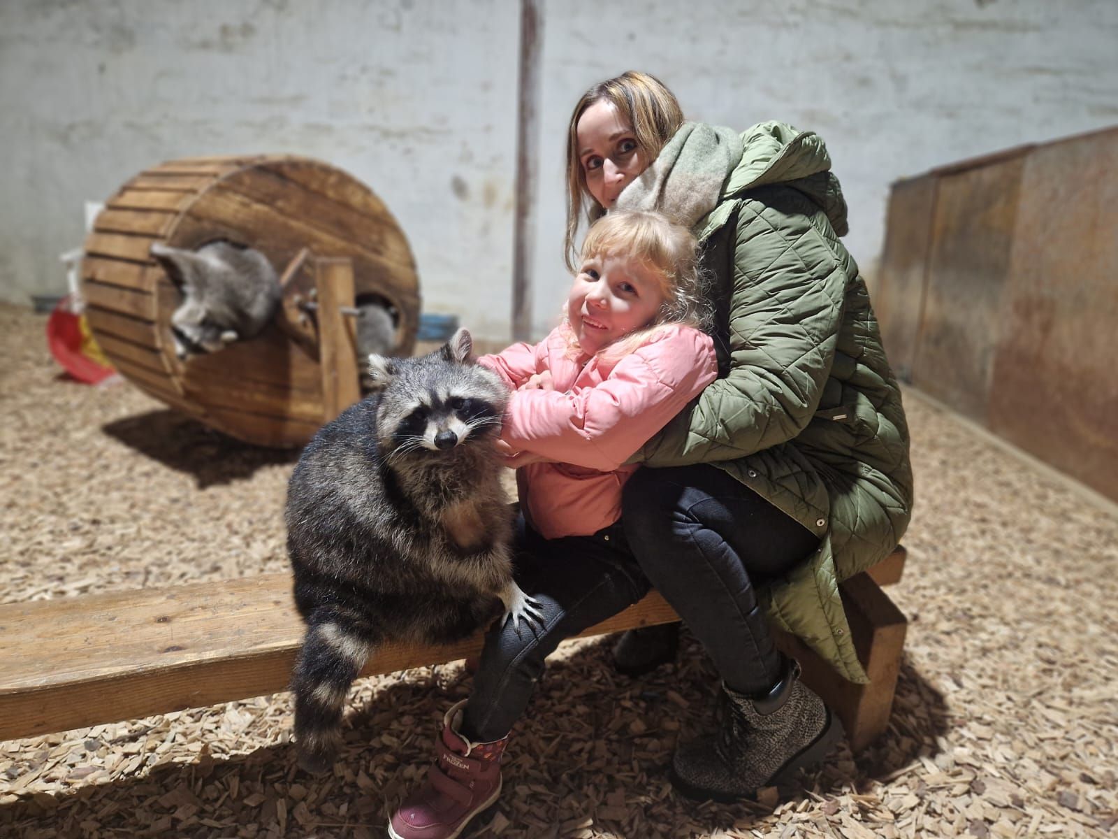 Eine Frau und ein kleines Mädchen sitzen mit einem Waschbären auf einer Bank.
