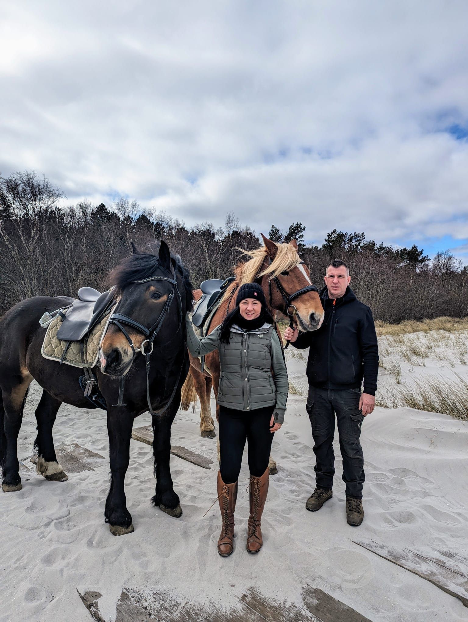 Ein Mann und eine Frau stehen neben zwei Pferden an einem Sandstrand.