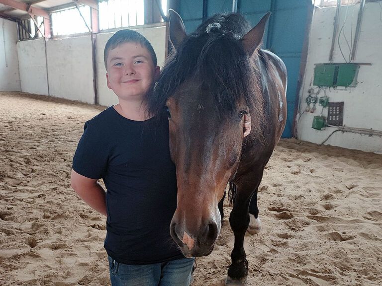 Ein kleiner Junge steht neben einem braunen Pferd in einem Stall.