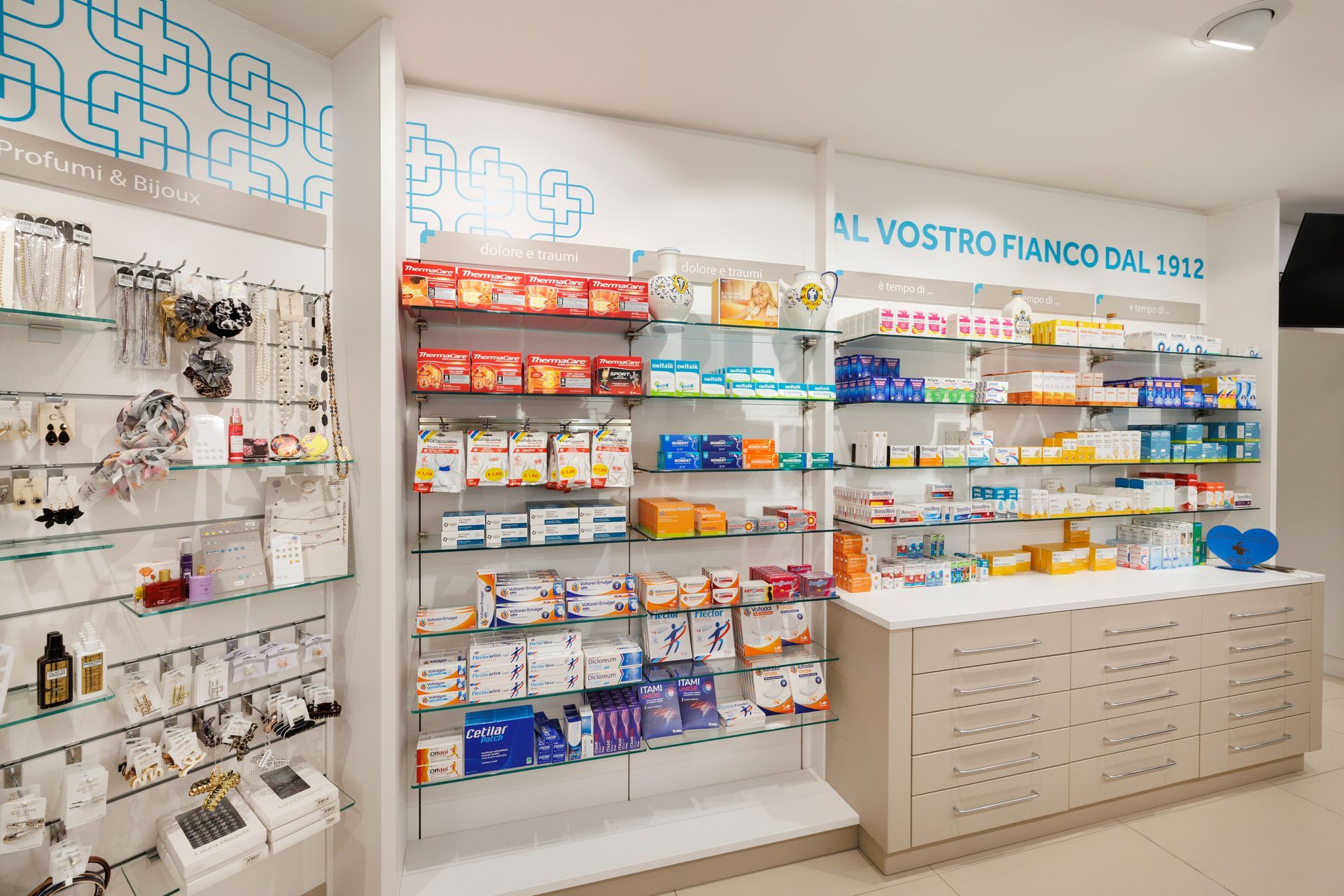 Interno luminoso della farmacia con scaffali pieni di medicinali e un bancone bianco espositivo.