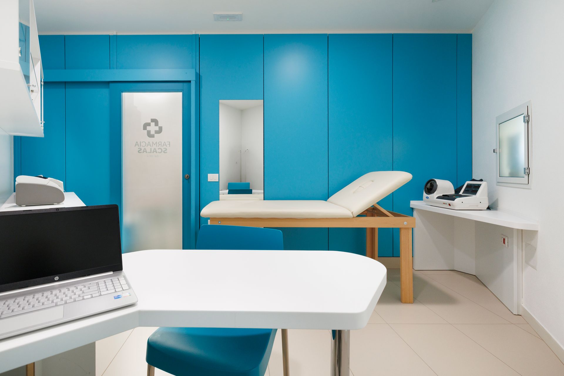 Luminosa sala per visite mediche con armadietti blu, lettino da visita, scrivania e attrezzature cliniche.