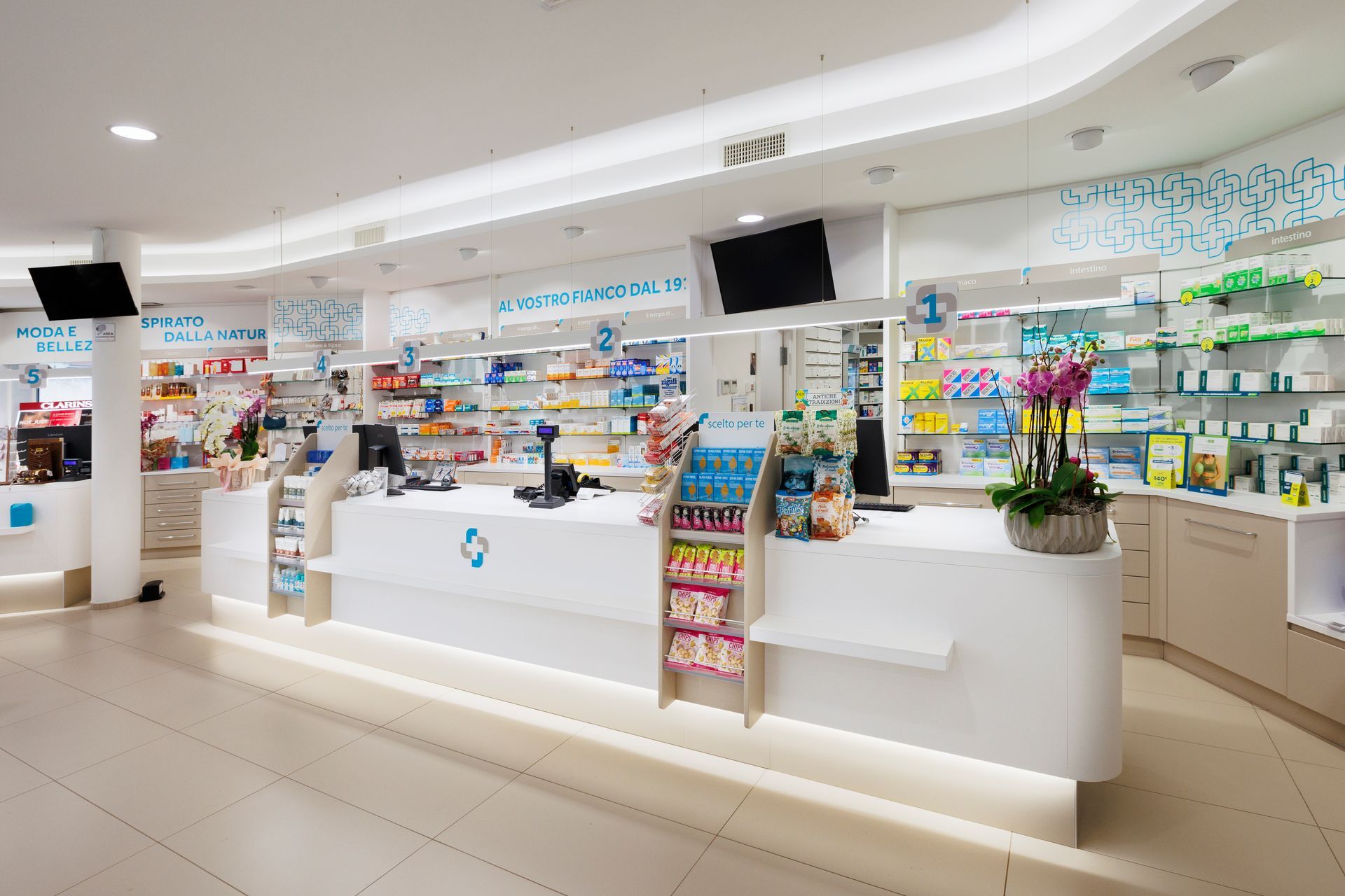 Luminoso interno di farmacia con banconi bianchi, scaffali di medicinali e colorati espositori di prodotti.