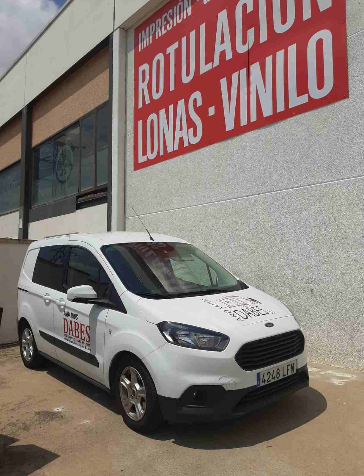 Furgoneta blanca aparcada frente a una tienda de rótulos con un letrero rojo que dice “ROTULACION LONAS VINILO” en la pared