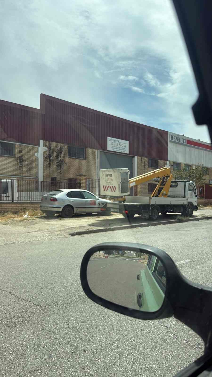 Vista de una nave industrial desde la calle, con coches aparcados y un camión de servicios bajo un cielo nublado