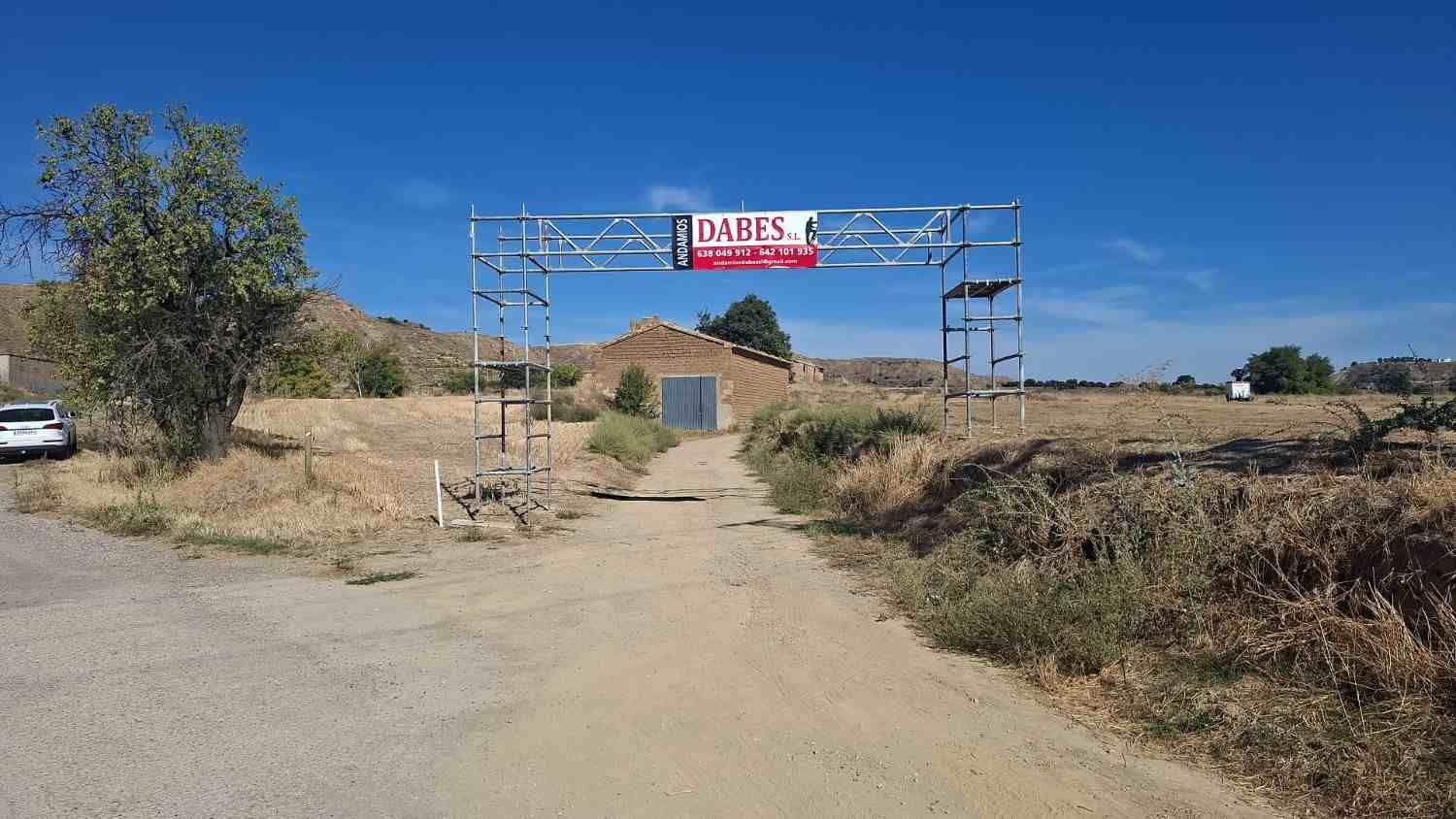 Camino de tierra que lleva a una entrada con portón y un letrero, en un paisaje seco y cubierto de hierba bajo un cielo azul