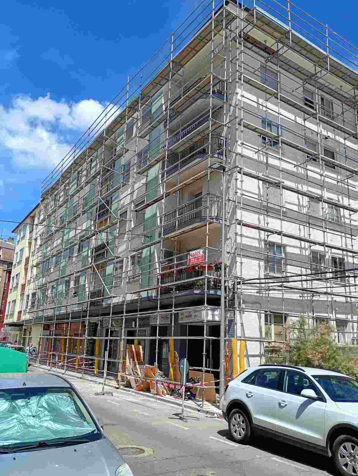 Edificio de apartamentos en obras, cubierto de andamios, en una calle soleada con coches aparcados