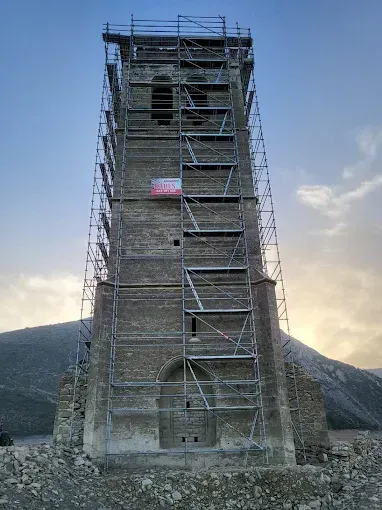 Torre de piedra rodeada de andamios y en restauración, con montañas al fondo al anochecer