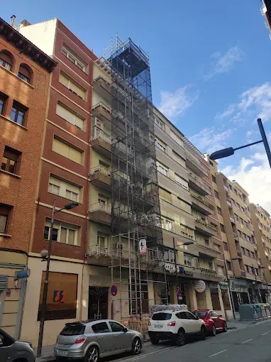 Calle urbana con edificios de apartamentos, coches aparcados y andamios en una fachada bajo un cielo azul