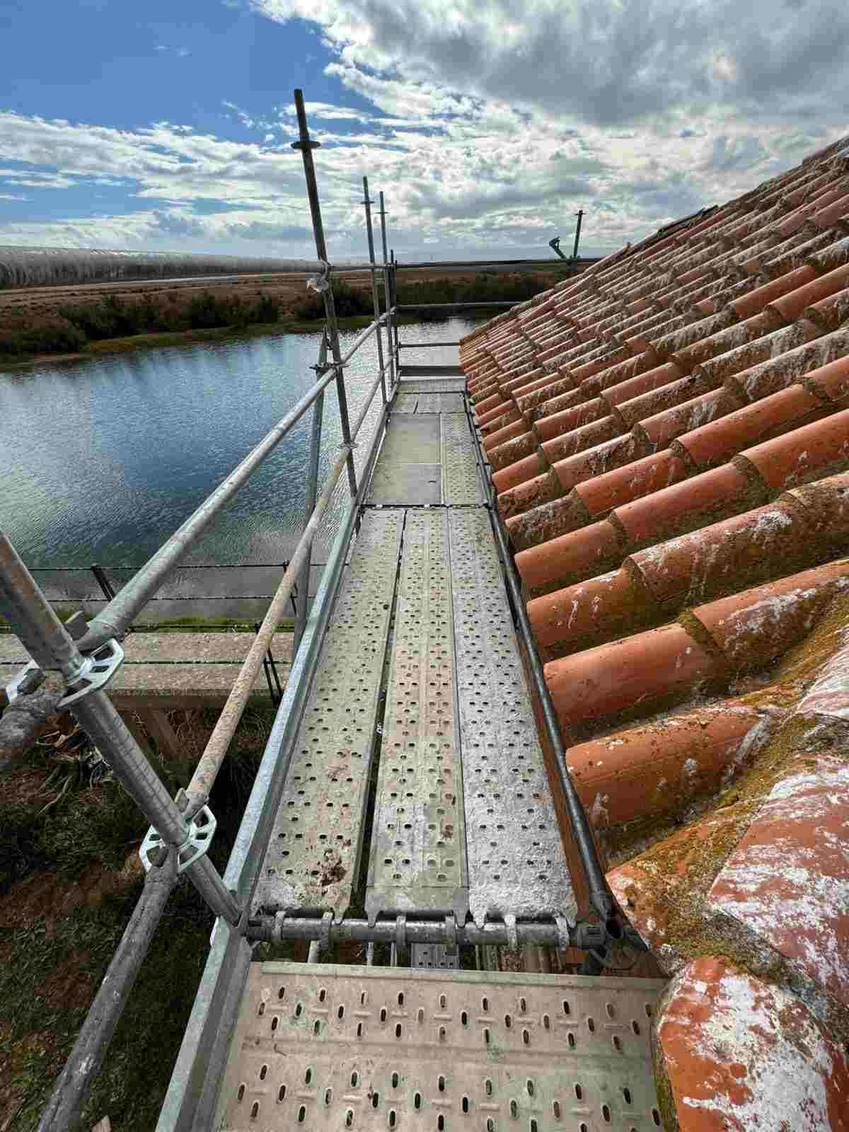 Pasarela estrecha de andamio junto a un tejado de tejas rojas sobre un canal, bajo un cielo nublado