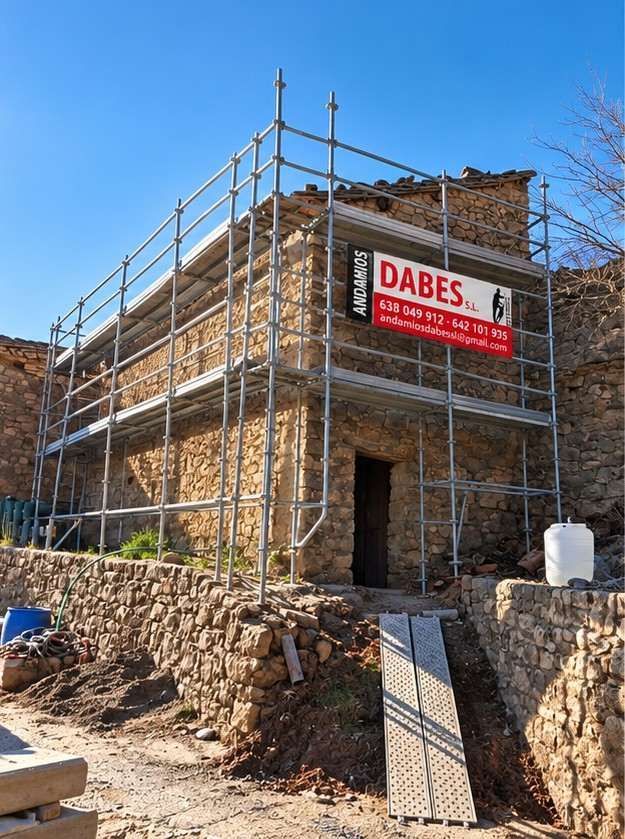Edificio de piedra en construcción, con andamios metálicos y un letrero rojo de DABES bajo un cielo azul
