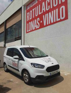 Furgoneta blanca de reparto aparcada frente a una tienda con un letrero rojo que dice «ROTULACIÓN LONAS VINILO»
