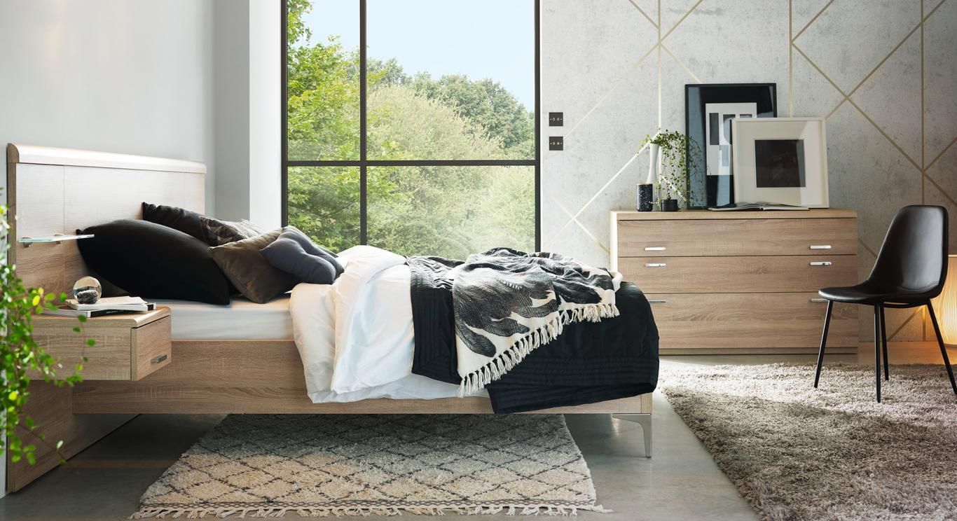 Chambre avec un lit en bois, une commode et une chaise noire ; grande fenêtre donnant sur des arbres.