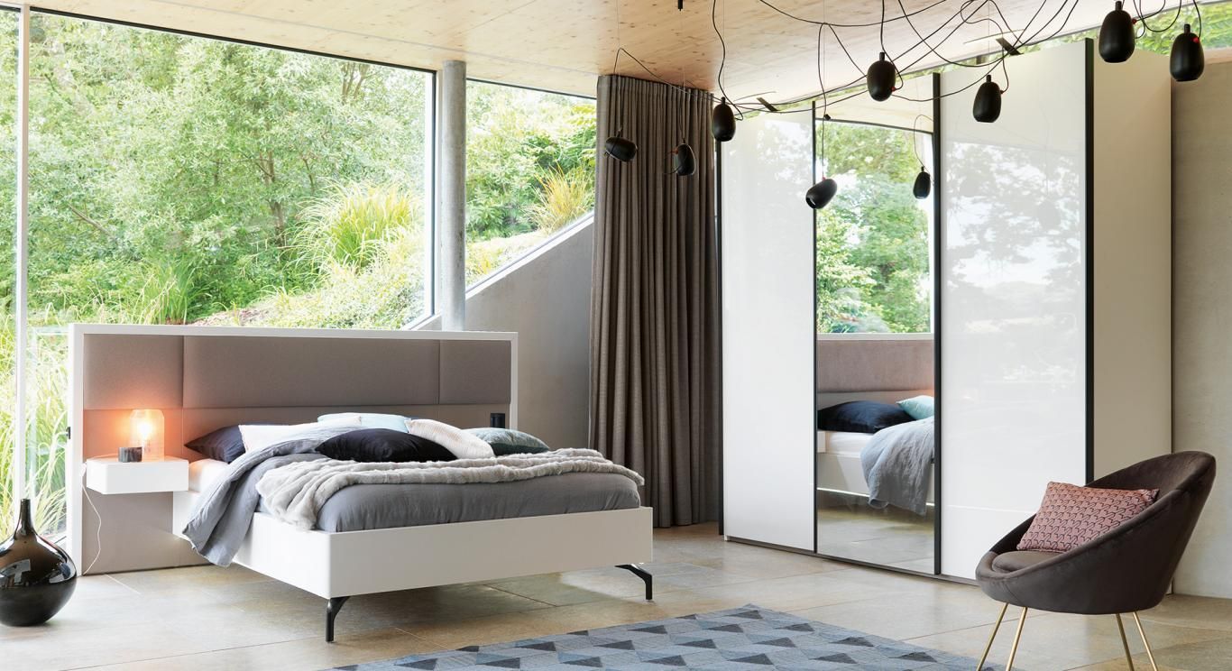 Chambre moderne avec de grandes fenêtres, un lit, une armoire à miroir et un fauteuil.