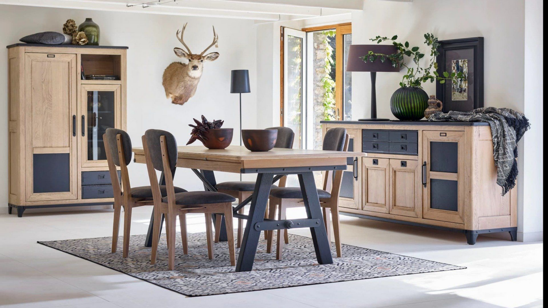 Salle à manger rustique avec mobilier en bois, comprenant une table et des chaises, un buffet et une armoire. Décoration murale en forme de tête de cerf.