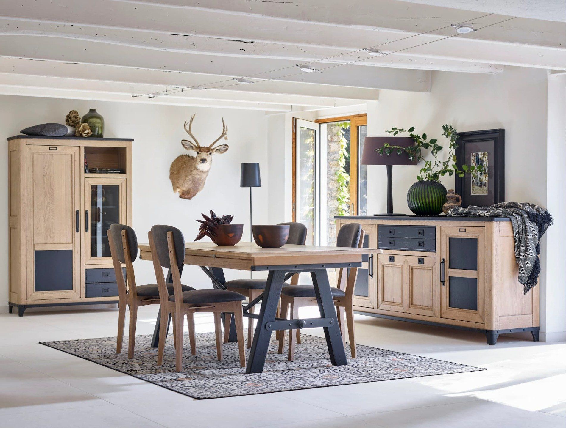 Salle à manger avec mobilier en bois, comprenant une table, des chaises et des armoires, ornée d'une tête de cerf et d'objets décoratifs.