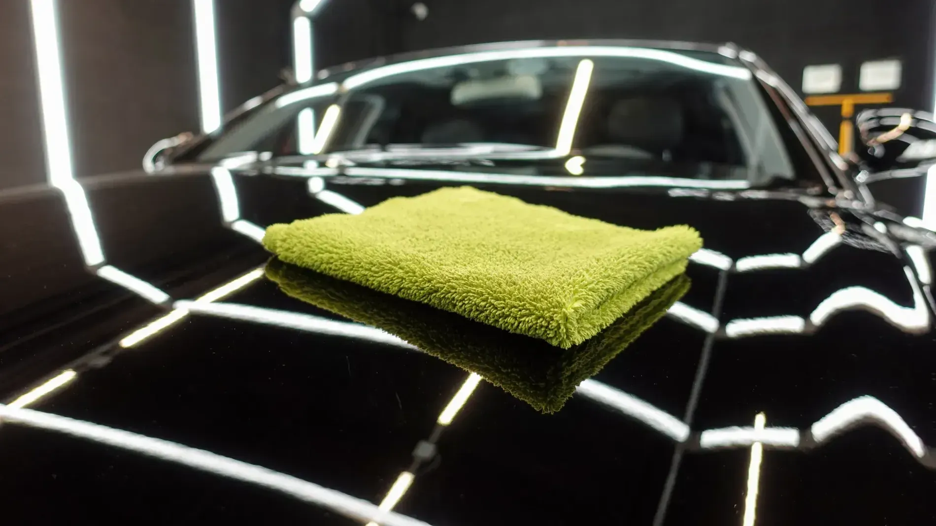 Paño de microfibra verde sobre el capó de un automóvil negro brillante, que refleja las brillantes luces del techo.