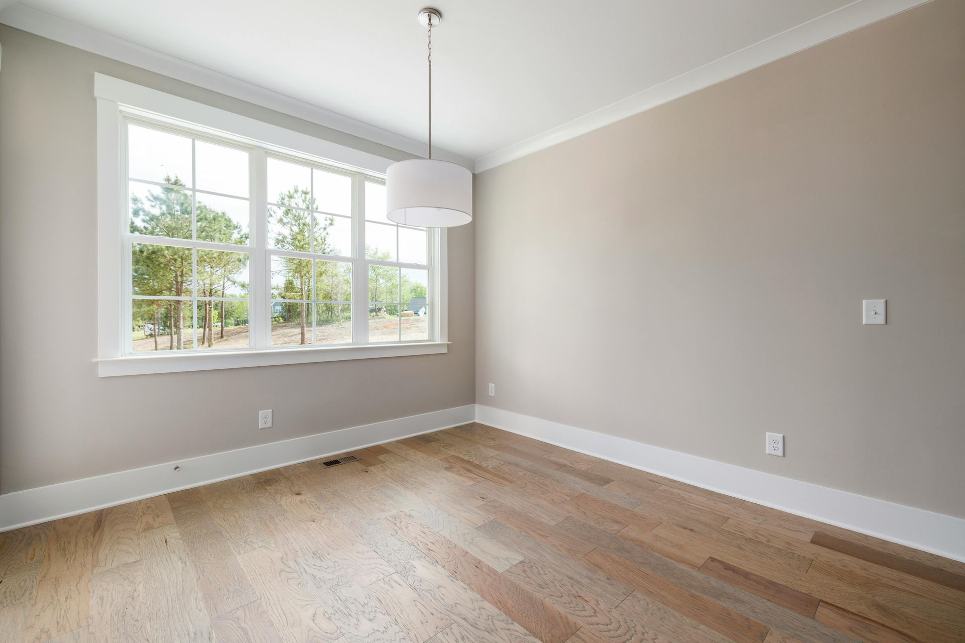 Tyhjä huone, jossa on suuri ikkuna ja puulattia. Neutraalit seinät, valkoiset listat ja riippuvalaisin.